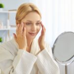 Young blonde woman in bathrobe gently applying skincare cream to her face using fingertips, looks into mirror touching face, thinks about plastic surgery or blepharoplasty. Skincare procedures concept