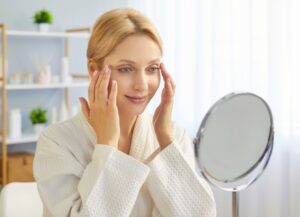 Young blonde woman in bathrobe gently applying skincare cream to her face using fingertips, looks into mirror touching face, thinks about plastic surgery or blepharoplasty. Skincare procedures concept